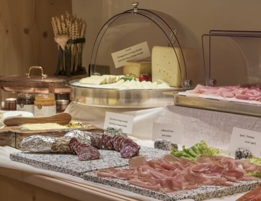 Close-up of a buffet table with cured meats, cheeses, and decorative dishes on a white cloth against a beige wall.
