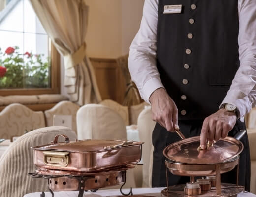 A man in a white shirt and black vest opens a copper serving dish in an elegant dining room with natural light.