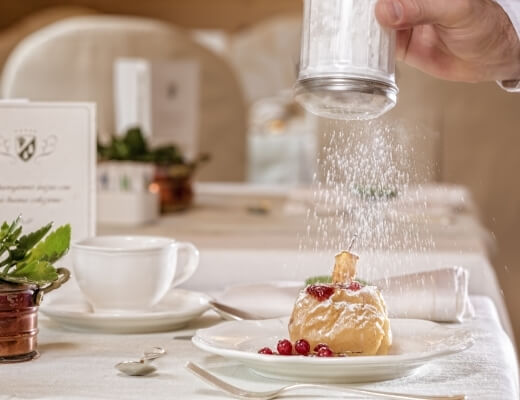 A hand sprinkles sugar on a cream puff dessert at a formal table setting with white plates and potted plants.