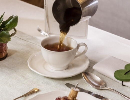 A white teacup on a saucer with tea being poured from a silver teapot, set on a white tablecloth with elegant tableware.