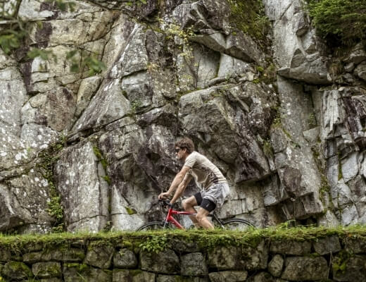 Mężczyzna na rowerze przed skalistym klifem, ubrany w jasny t-shirt i czarne szorty, jedzie na czerwonym rowerze.