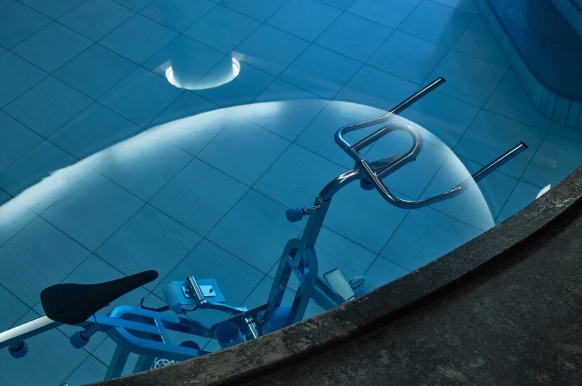 Ein blauer Trainingsfahrrad steht in einem Pool mit blauen Fliesen, umgeben von einem Betonrand und einem runden Licht.