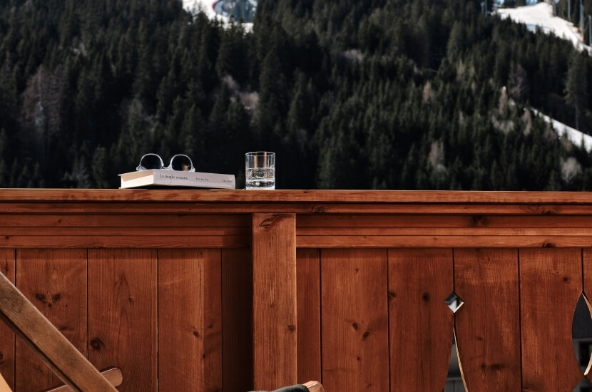 Railing in legno con un libro, occhiali da sole e un bicchiere d'acqua, sullo sfondo di una montagna innevata.