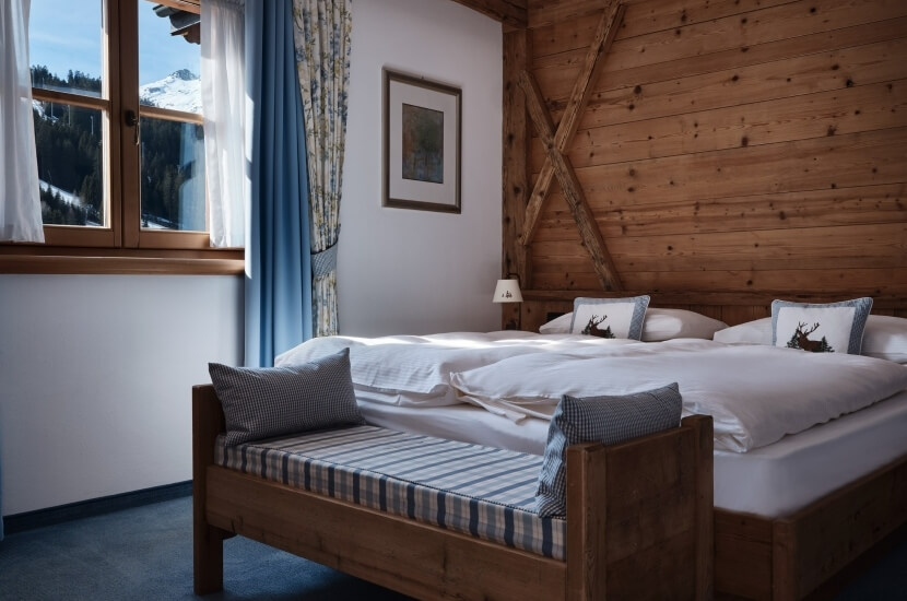 Serene rustic bedroom with two beds, cozy bench, nightstand, and mountain view through a window, exuding tranquility.