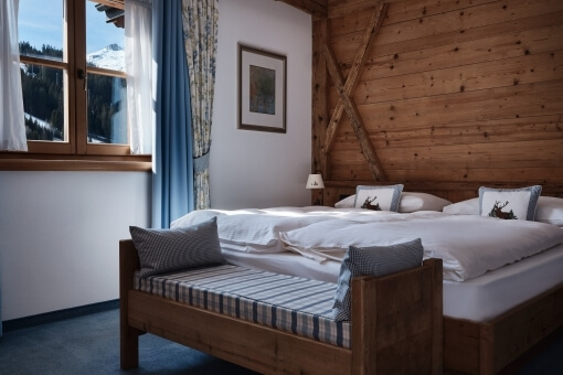 Dimly lit bedroom with a large bed, striped bench, white walls, blue carpet, and a window with curtains.
