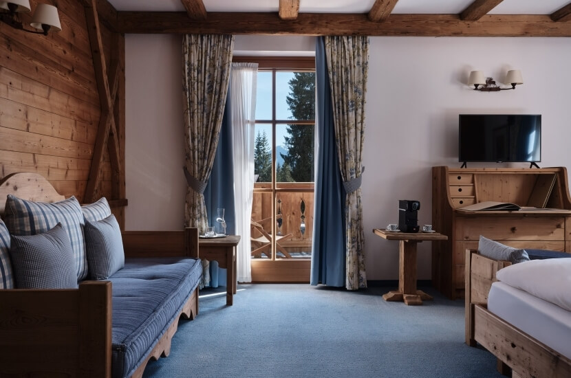 Cozy room with blue couch, beds, TV, and large window with blue curtains, featuring rustic beams and blue carpet.