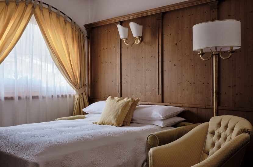 Serene bedroom with a bed, yellow chairs, lamp, and sunlight through tied-back yellow curtains, creating a calm atmosphere.