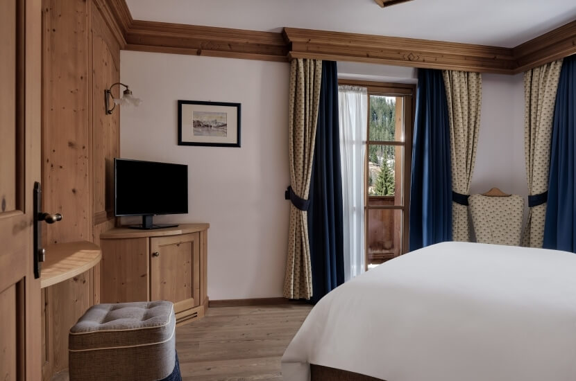Camera da letto rustica con letto, TV, finestra e ottomano, decorata in toni caldi e vista su alberi all'esterno.