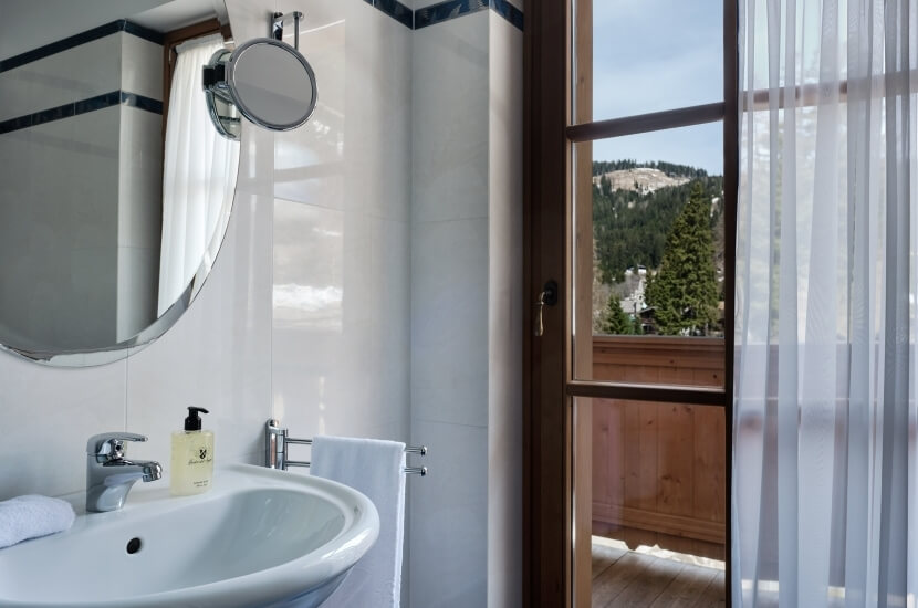 Bagno elegante con lavabo bianco, specchio ovale, finestra con vista su paesaggio montano e pavimento in legno chiaro.