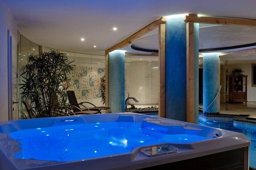 Ein Whirlpool mit blauem Wasser und Lichtern, umgeben von Pflanzen, Säulen und einer Treppe in einem entspannenden Spa-Ambiente.