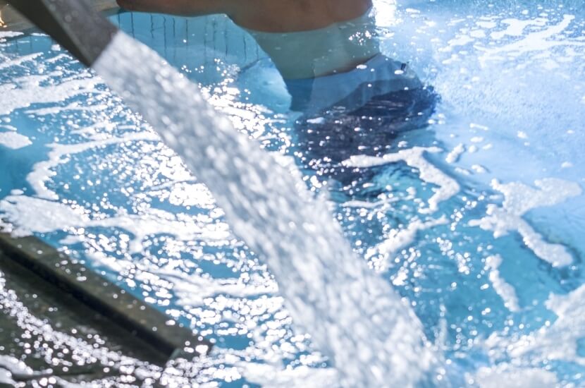 Personenbeine im Pool mit spritzendem Wasser, hellblaue Badehose, dunkelblaue Schwimmschuhe, sonnige, erfrischende Atmosphäre.
