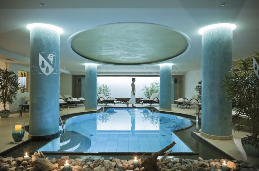 Innenpool mit Wassermerkmal, umgeben von Pflanzen, Liegen und natürlichem Licht in einem luxuriösen Spa-Ambiente.