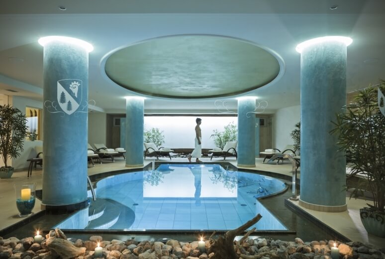 Luxuriöser Innenpoolbereich mit einem Mann, umgeben von Pflanzen, Kerzen und einem Wassermerkmal, strahlt Ruhe und Eleganz aus.