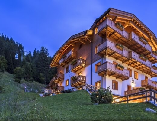 Edificio bianco con accenti in legno marrone su una collina, circondato da alberi e un cielo blu, con una funivia a sinistra.