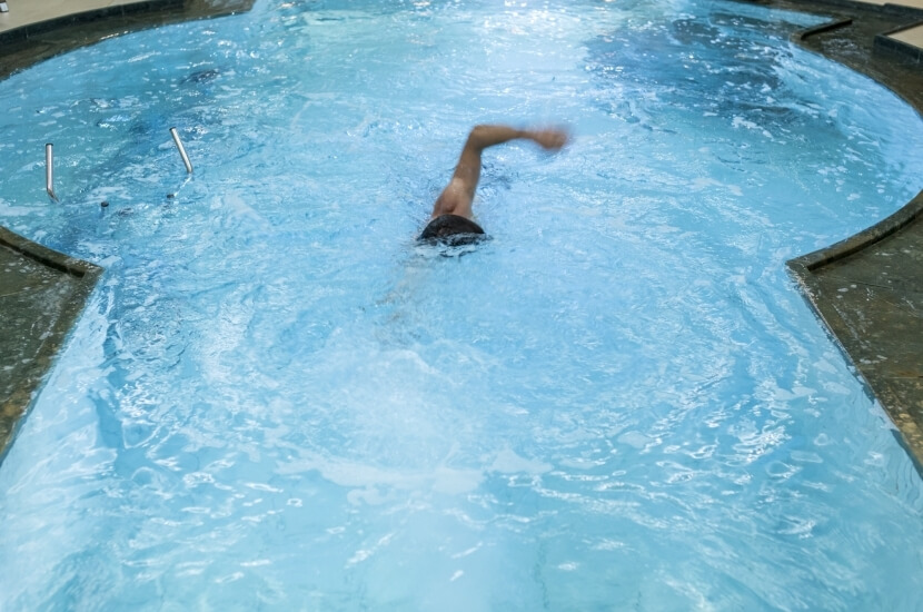 Eine Person schwimmt vertikal im Pool, trägt schwarze Badehosen und hat einen Arm über Wasser, während der andere untergetaucht ist.