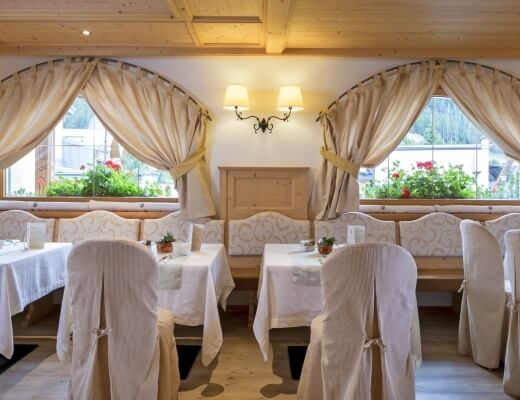A well-lit dining room with white tablecloths, potted plants, and exposed wooden beams, featuring two windows and a doorway.