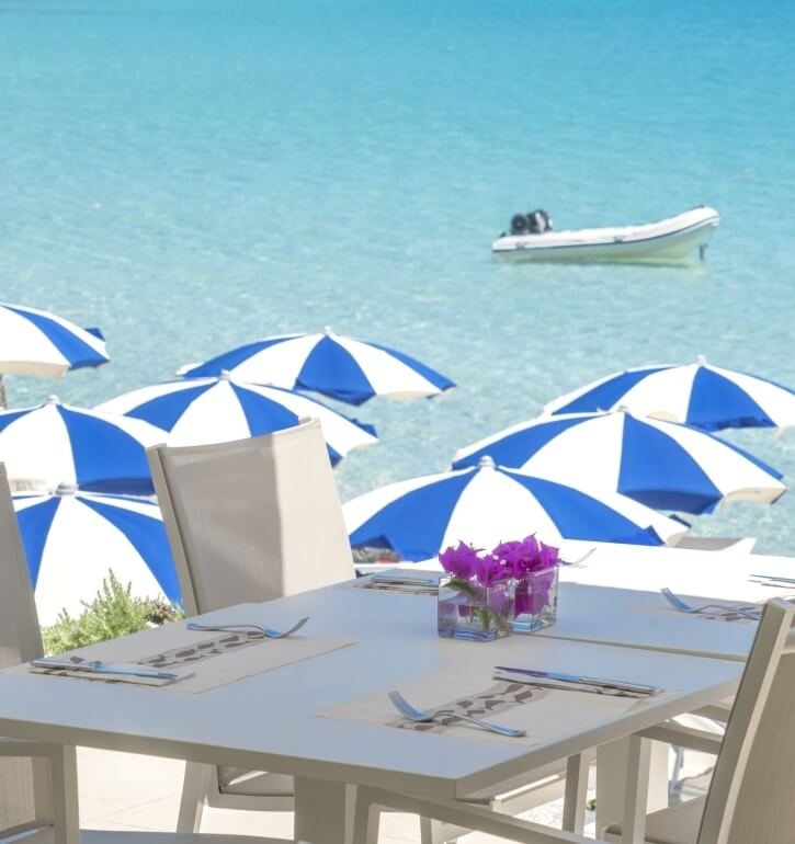 Tisch mit Platzdeckchen und Blumen, Blick auf türkisfarbenes Wasser.