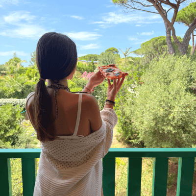 Persona con pizza su un balcone verde, circondata da alberi e cielo blu.