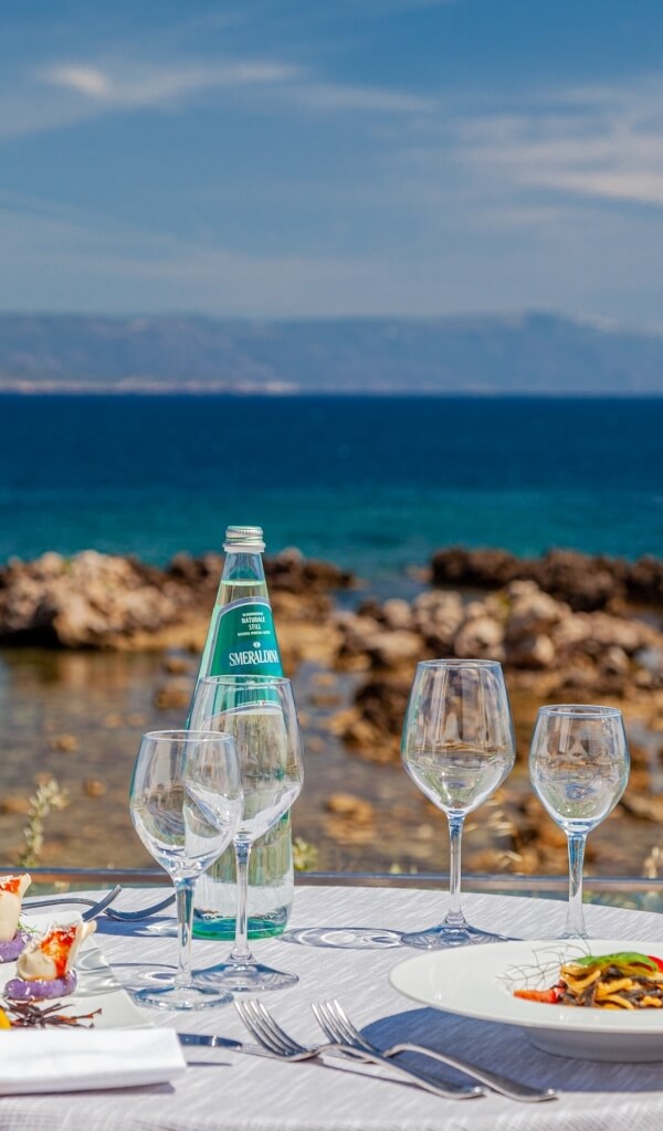 Tavolo all'aperto con cibo, vino e vista su acqua e colline.