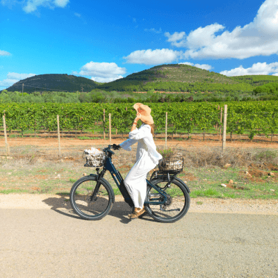 Persona in abito bianco su bici elettrica con cesti, vigneto sullo sfondo.