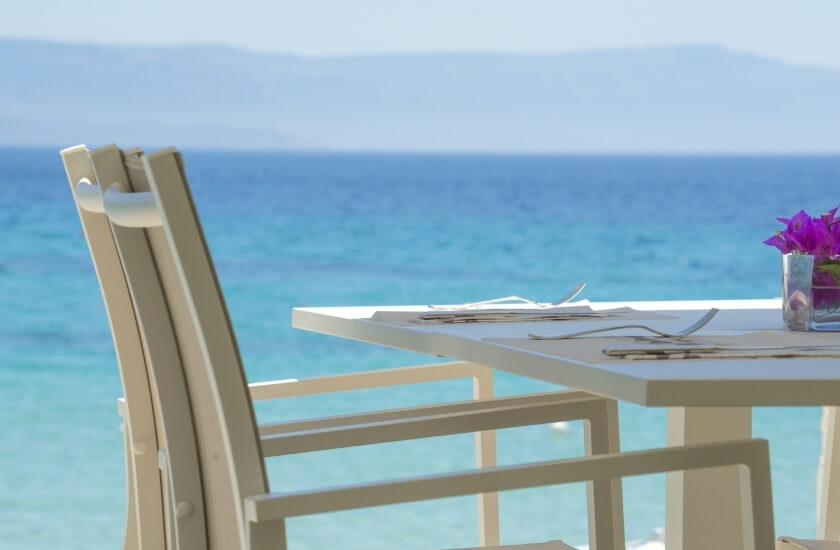 Tavolo bianco con posate, vaso di fiori viola e oceano blu.