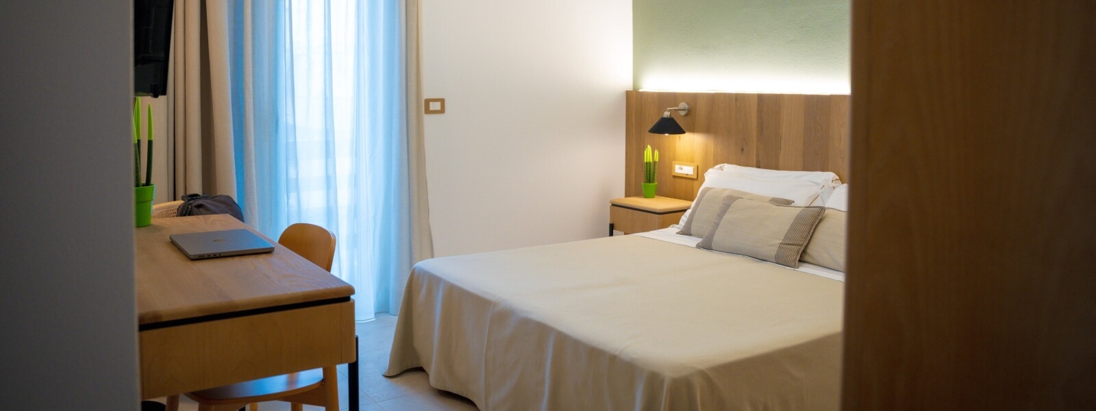 Camera d'hotel con letto, scrivania, piante e tende blu.