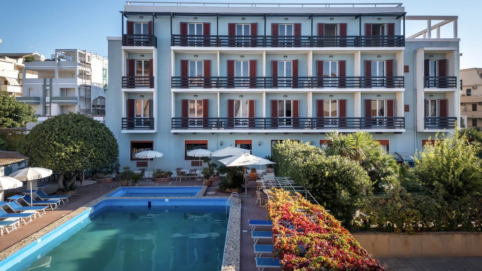 Hotel con facciata azzurra, piscina e aree verdi circostanti.