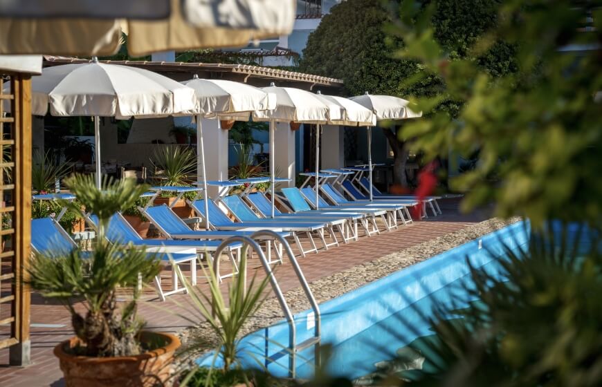 Area piscina con sedie a sdraio blu, ombrelloni bianchi e piante.