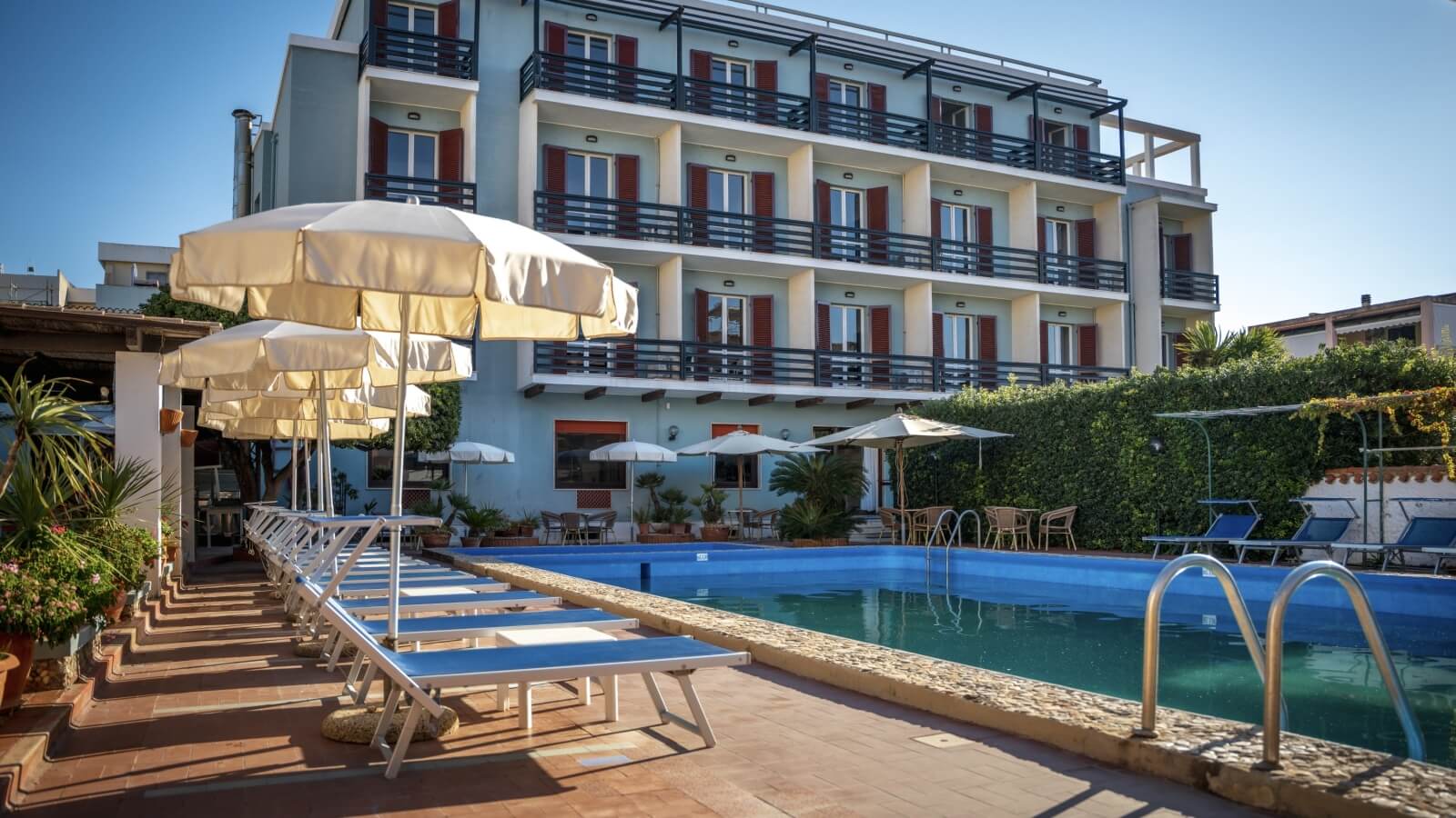 Piscina all'aperto con sdraio, ombrelloni e edificio blu sullo sfondo.