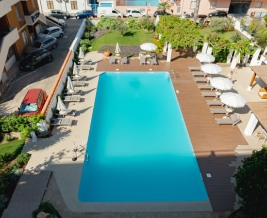 Piscina rettangolare con deck in legno, sdraio e auto parcheggiate.