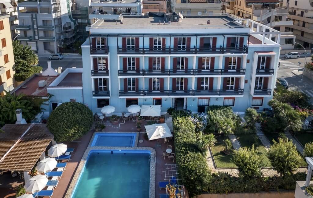 Hotel with pool, lounge chairs, and green spaces; light blue building.   Hotel con piscina, lettini e spazi verdi; edificio azzurro.