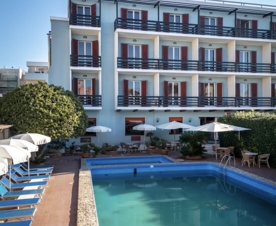 Hotel with balconies, pool, lounge chairs, and a large tree.   Hotel con balconi, piscina, lettini e un grande albero.