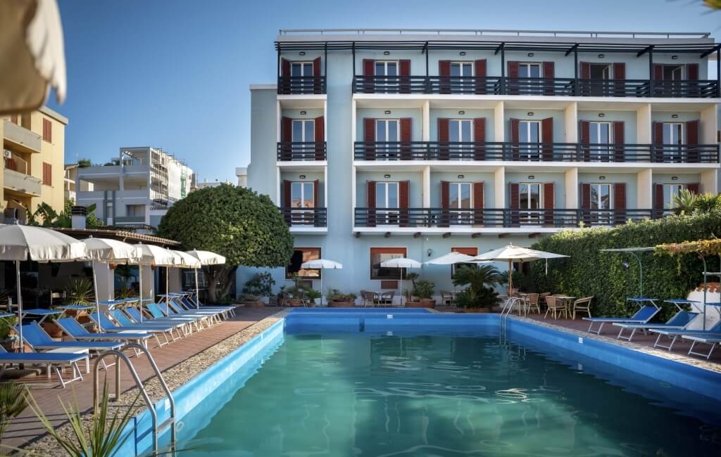 Piscina con lettini e ombrelloni, edificio blu sullo sfondo.