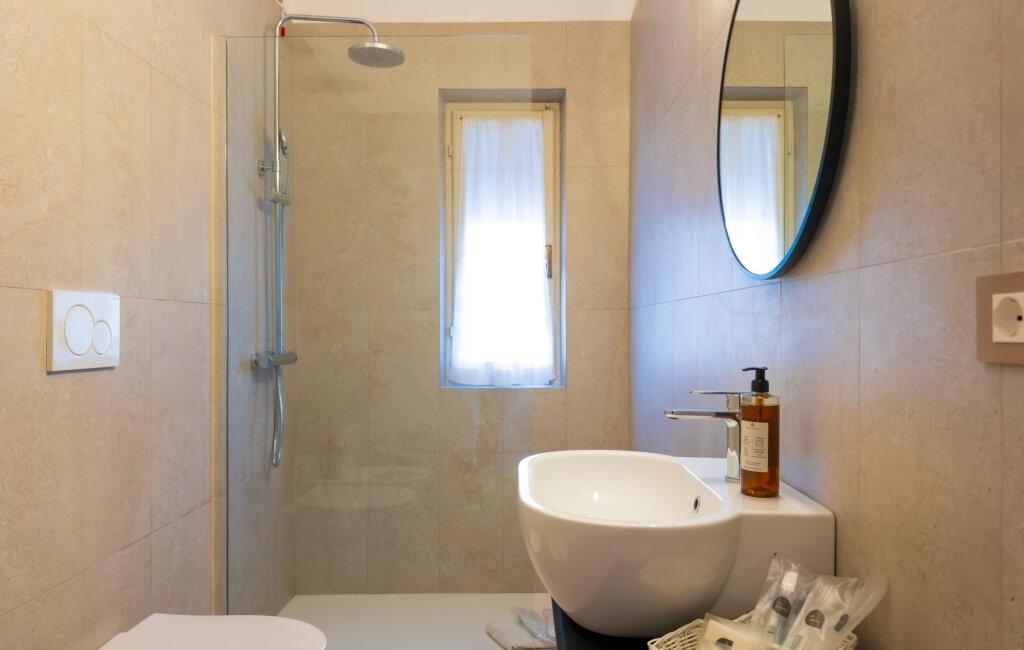 Bagno con doccia in vetro, lavabo, specchio rotondo e finestra.