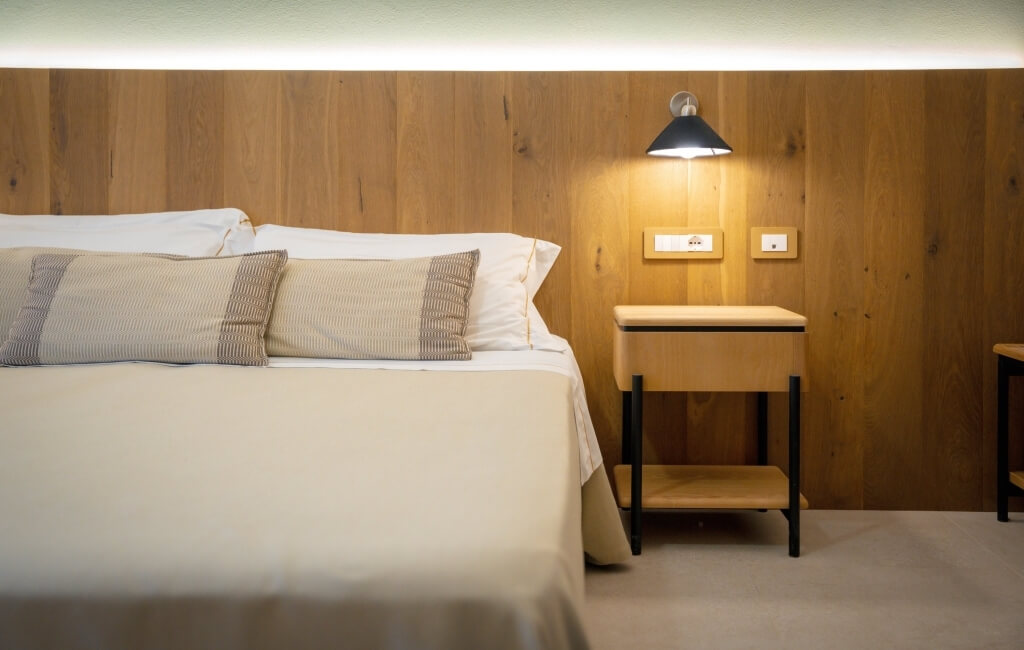 Letto con coperte beige, testiera in legno e lampada da comodino.