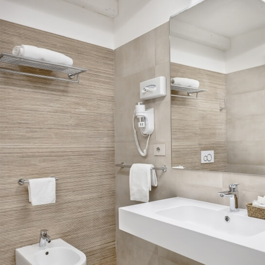 Bagno moderno con lavabo bianco, asciugamani, asciugacapelli e piastrelle chiare. Atmosfera pulita e ordinata.