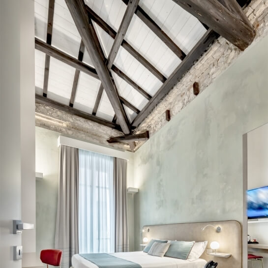 Camera moderna e serena con travi a vista, letto bianco, sedia rossa e TV montata su parete.