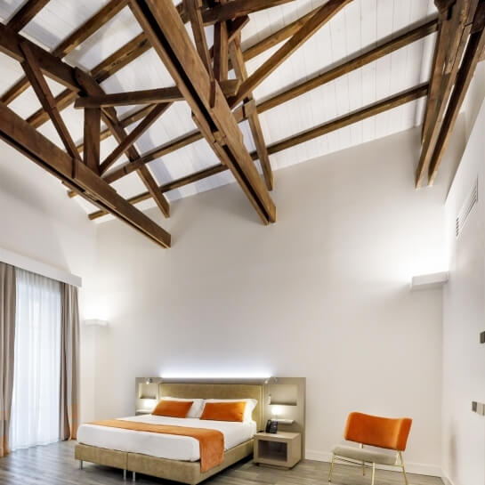 Modern minimalist bedroom with white walls, wooden beams, large bed with orange blanket, nightstand, and natural light.