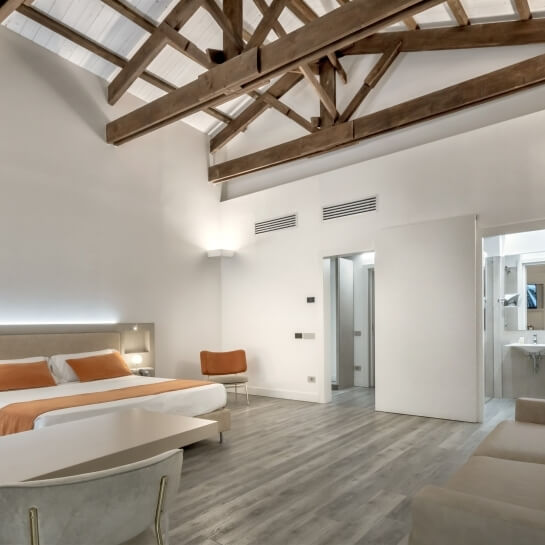 Modern hotel room with high ceiling, exposed beams, bed with orange throw, nightstand, chair, and visible bathroom doorway.