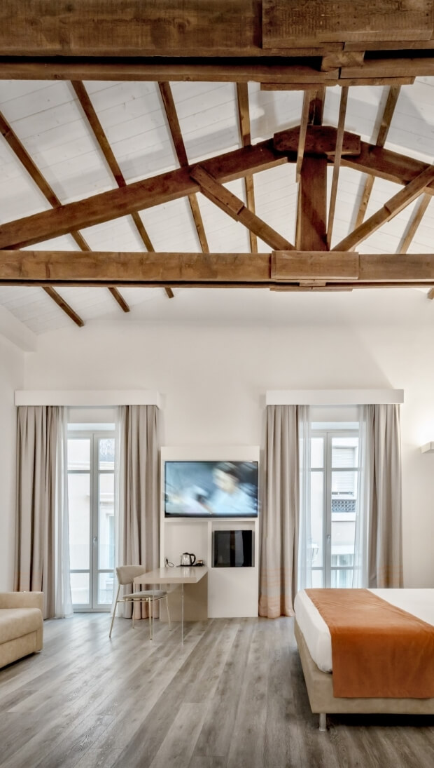Spazio abitativo moderno e minimalista con letto, divano colorato, finestre luminose e travi in legno a vista.