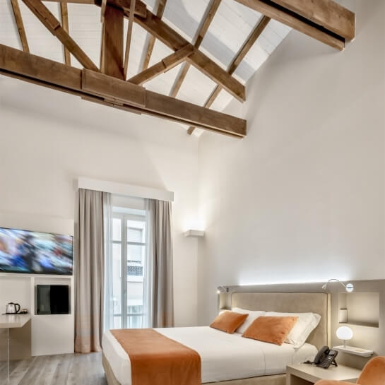 Modern white bedroom with a large bed, orange pillows, television, wooden beam, and serene atmosphere.