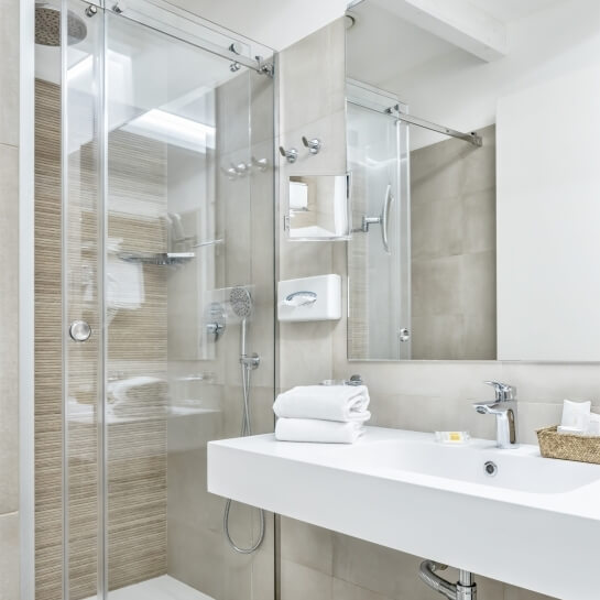 Modern all-white bathroom with glass shower, white towels, sink, and light-brown tiled walls, exuding serenity and minimalism.