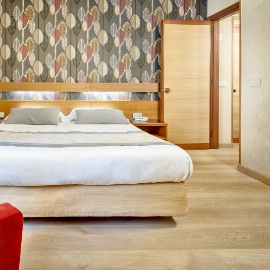Camera da letto serena con letto grande, parete decorativa a foglie, comodino con sveglia e libro, pavimento in legno chiaro.