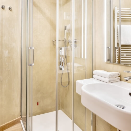 Bagno con doccia in vetro, lavabo bianco, specchi, asciugamani e piastrelle beige lucide.