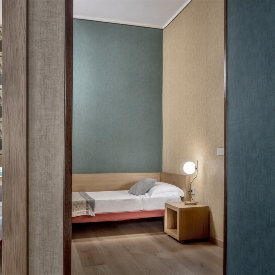 Una piccola stanza accogliente con un letto singolo rosso, comodino in legno e lampada, divisa da un muro verticale.