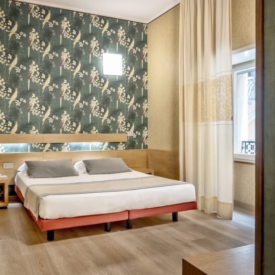 Camera da letto con due letti, finestra con tende bianche e carta da parati verde con fiori bianchi. Pavimento in legno chiaro.