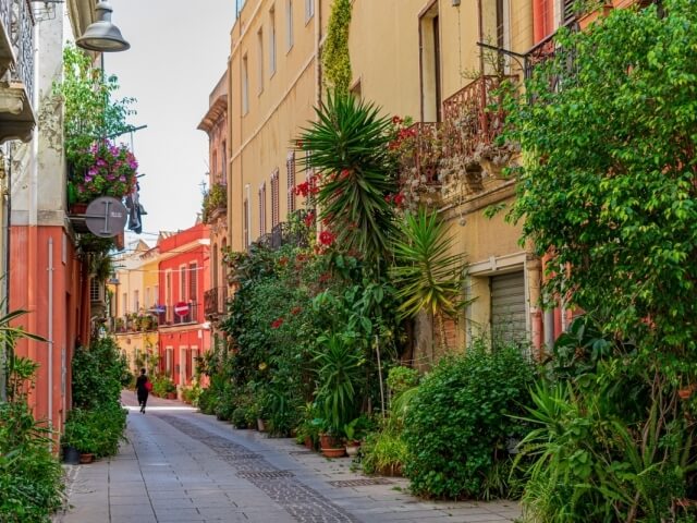 Una stretta strada di ciottoli con edifici color sabbia e rosso, piante verdi e una persona che cammina al centro.