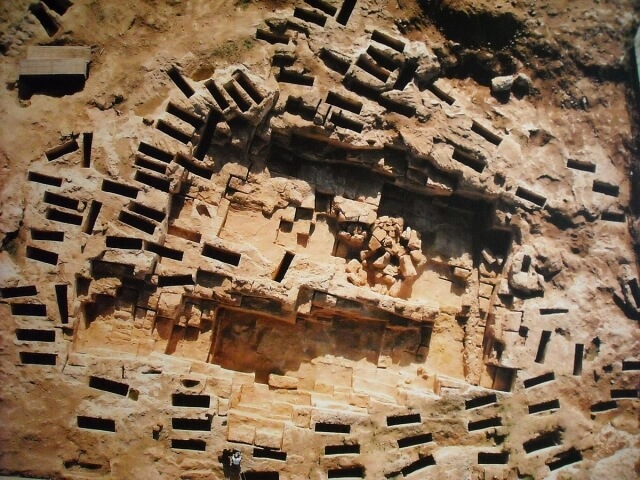 Struttura antica in rovina con mattoni di pietra, aperture rettangolari e terreno più chiaro circostante.