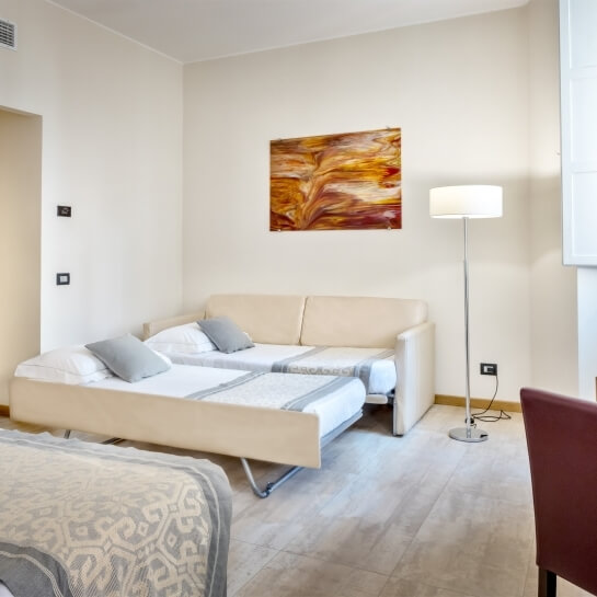 Una stanza con un divano letto, un dipinto colorato, una lampada e un'altra poltrona maroon, su pavimento chiaro.