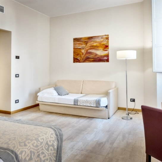 Camera minimalista con divano letto bianco, lampada, sedia rossa e opere d'arte, pavimento beige con striature grigie.